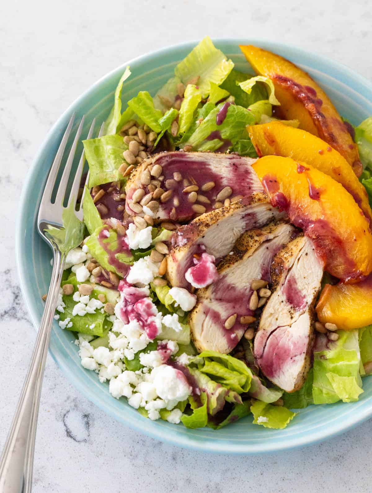 A leafy salad topped with sliced chicken, peach wedges, and crumbled goat cheese is drizzled with blueberry vinaigrette.