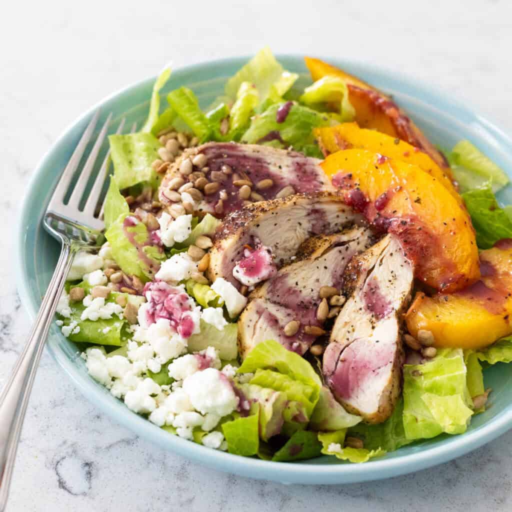 A leafy salad topped with sliced chicken, peach wedges, and crumbled goat cheese is drizzled with blueberry vinaigrette.