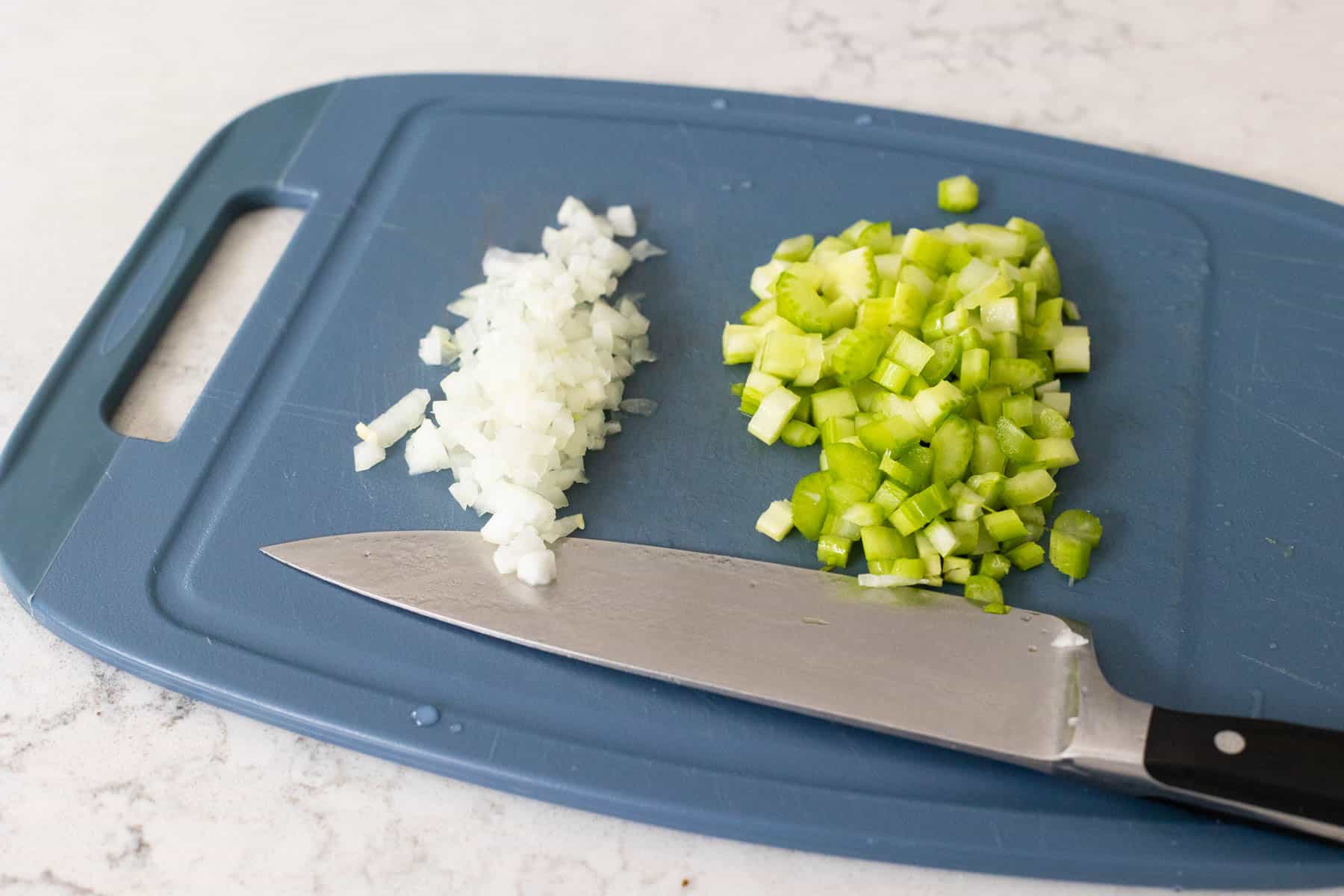 A blue cutting board with finely chopped onion on the left and diced celery on the right, accompanied by a sharp kitchen knife.