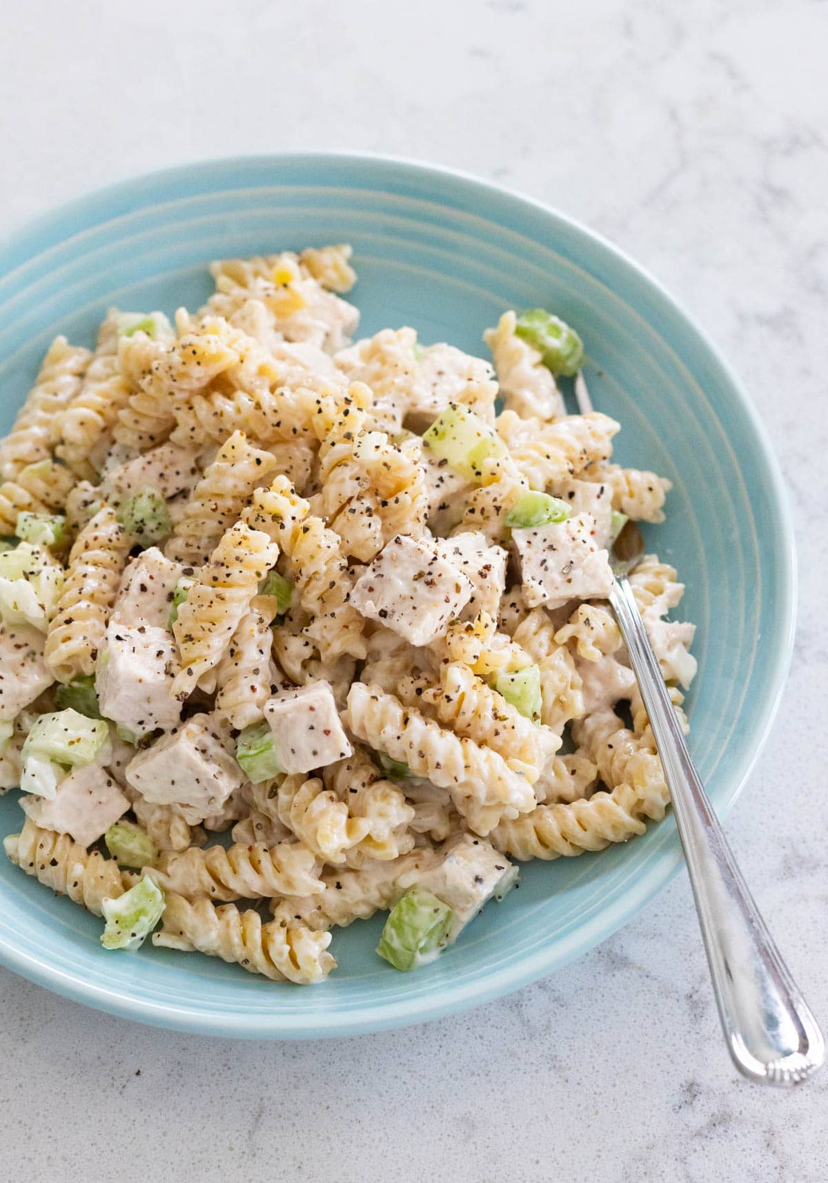 A bowl of chicken salad pasta salad with rotini pasta, chicken breast, celery and pepper, served on a blue plate with a fork.