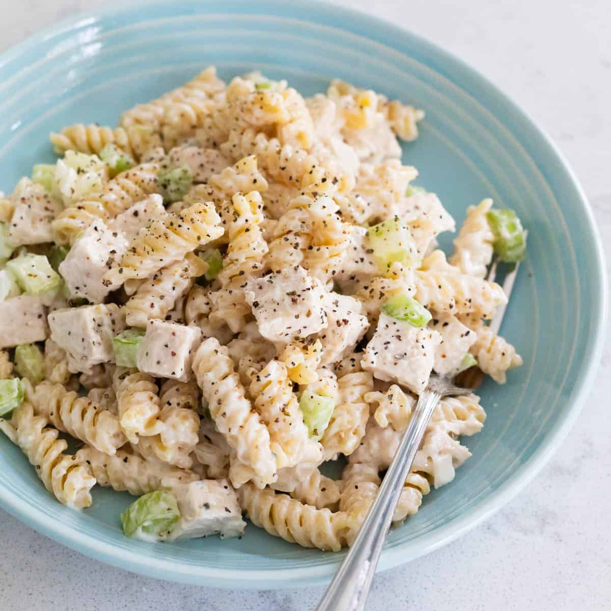 A blue bowl is filled with creamy pasta salad with chunks of chicken and chopped celery.