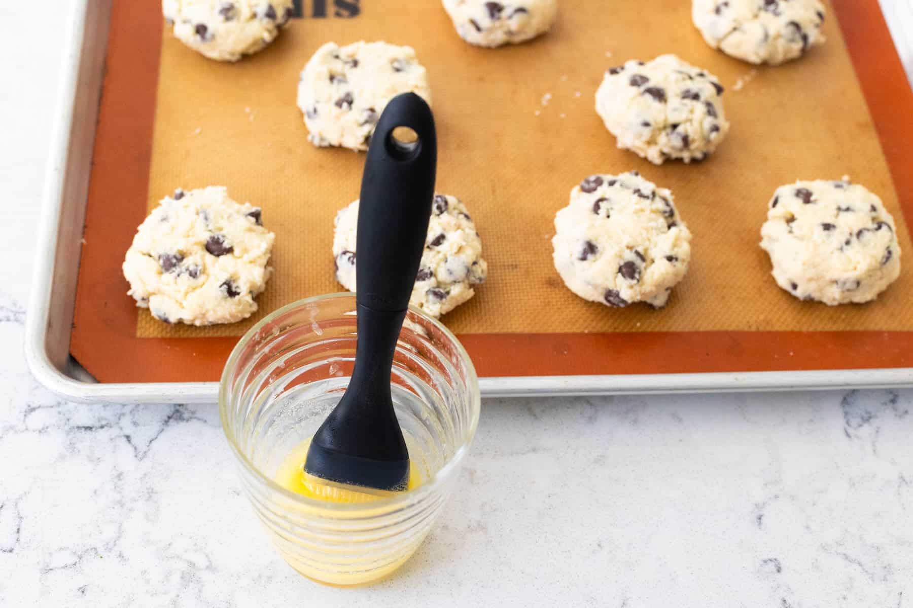 Unbaked chocolate chip scones on a baking sheet next to a glass of melted and a pastry brush.