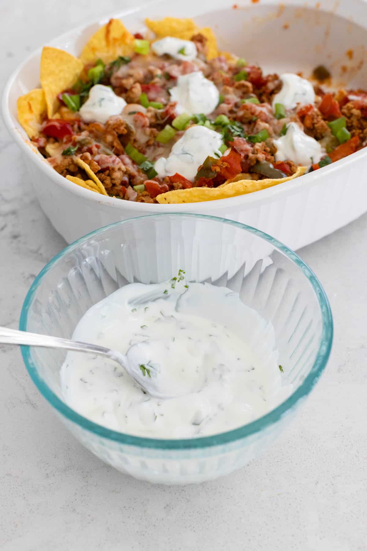 A bowl of cilantro lime crema with nacho casserole behind it.