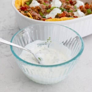 A bowl of cilantro lime crema has a spoon ready to dollop it over a nacho casserole that sits behind it.