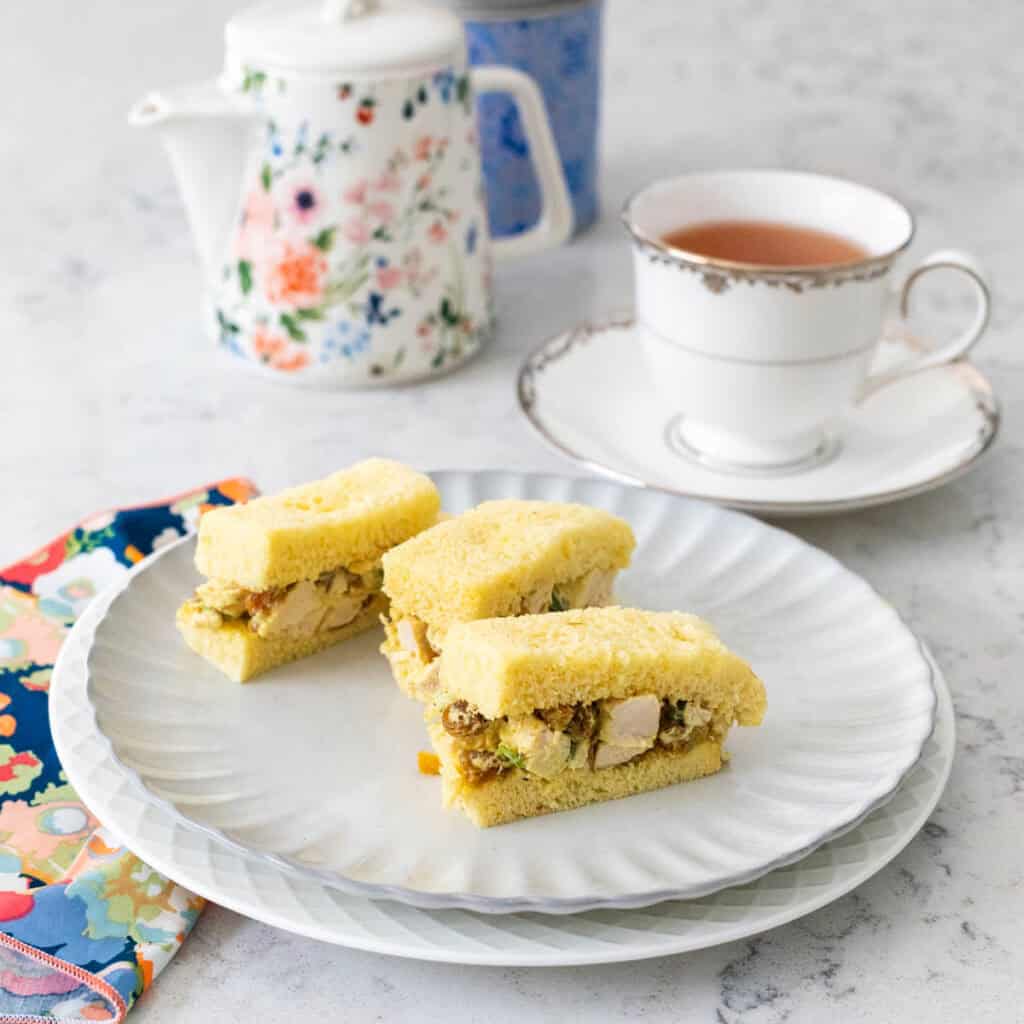A ruffled plate has 3 small coronation chicken finger sandwiches. Next to it sits a cup of Earl Grey tea and a little tea pot.
