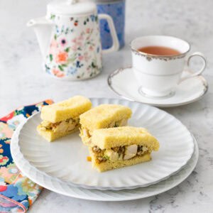 A ruffled plate has 3 small coronation chicken finger sandwiches. Next to it sits a cup of Earl Grey tea and a little tea pot.