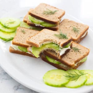 A white platter has several small bite-sized cucumber sandwiches with fresh dill. A bite has been taken out of the mini sandwich on top.