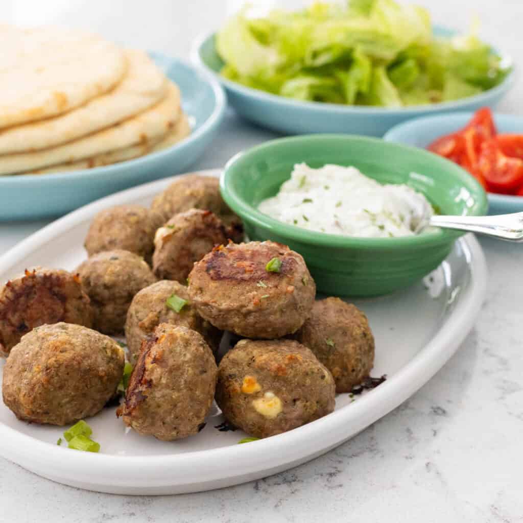 A platter of gyro meatballs is served with a bowl of tzatziki sauce. Pitas, lettuce, and chopped tomatoes are in the background.