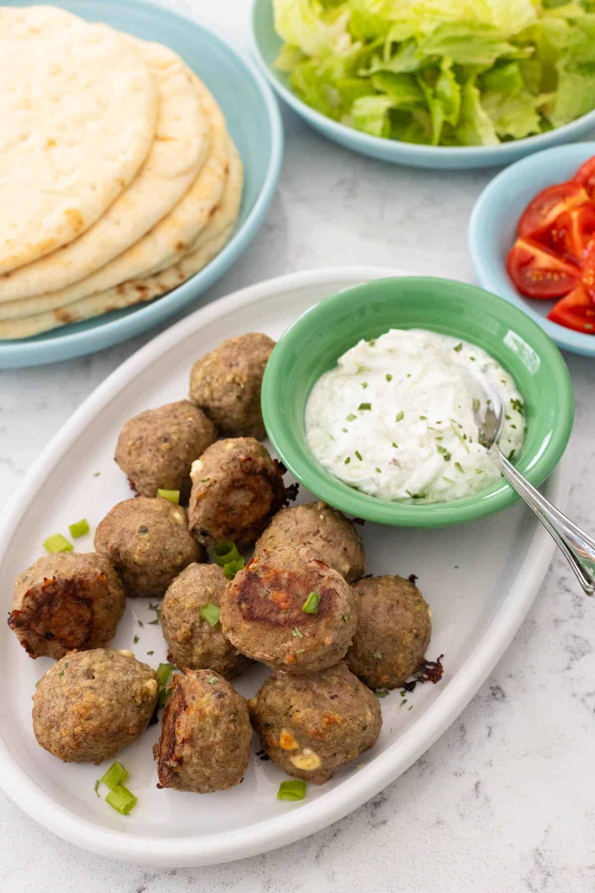 A platter of gyro meatballs is served with a bowl of tzatziki sauce. Pitas, lettuce, and chopped tomatoes are in the background.