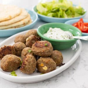 A platter of gyro meatballs is served with a bowl of tzatziki sauce. Pitas, lettuce, and chopped tomatoes are in the background.