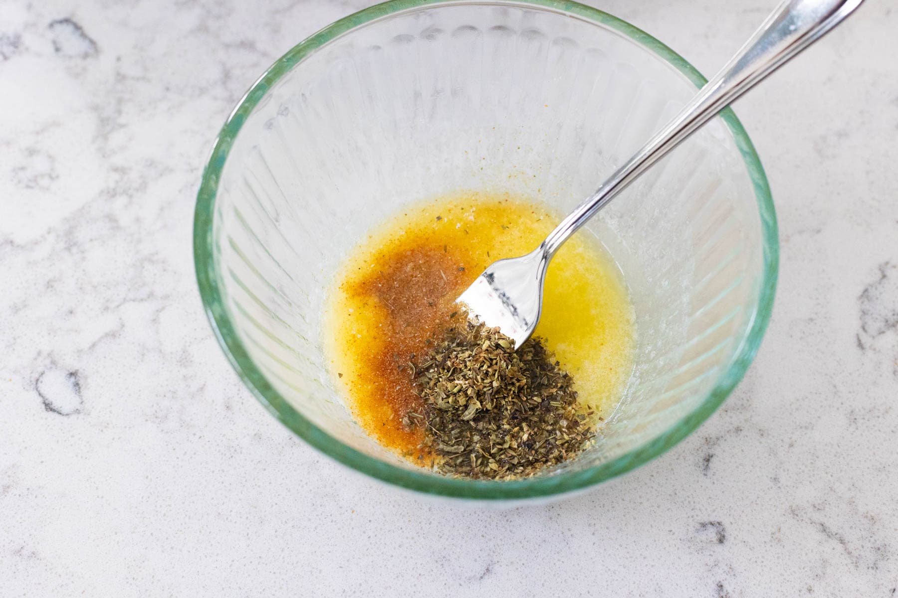 Melted butter mixed with herbs and spices in a glass bowl with a fork.