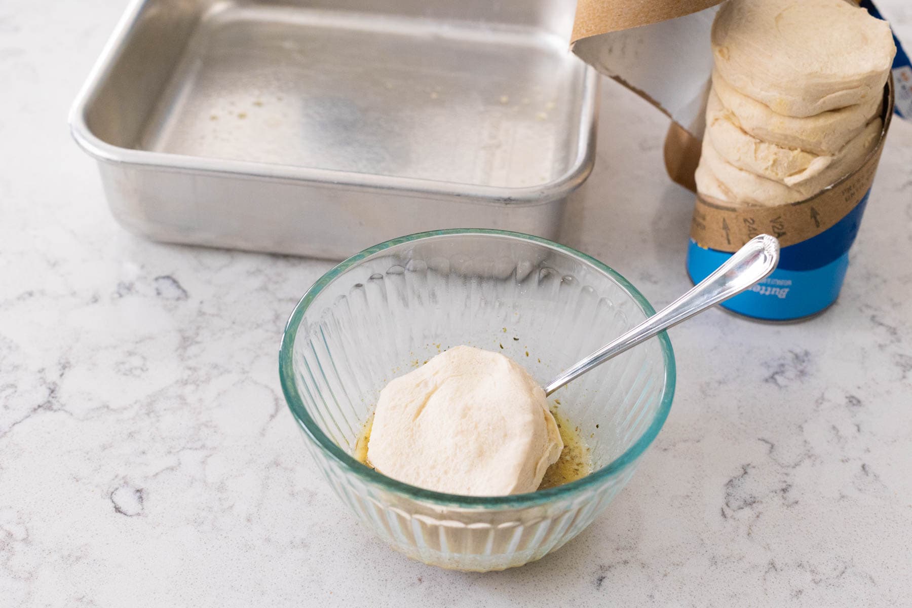 Unbaked biscuit being dipped into herb butter mixture in a glass bowl, with a baking pan and biscuit can nearby.
