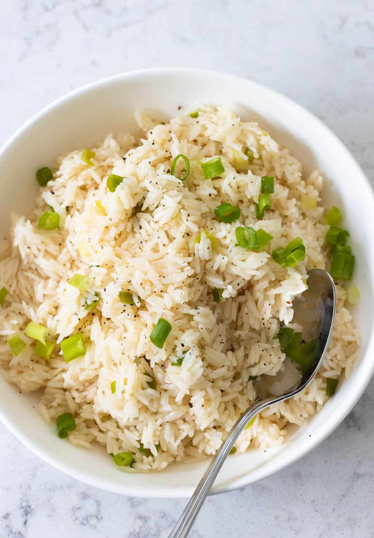 A bowl of cooked white rice garnished with chopped green onions served with a spoon.