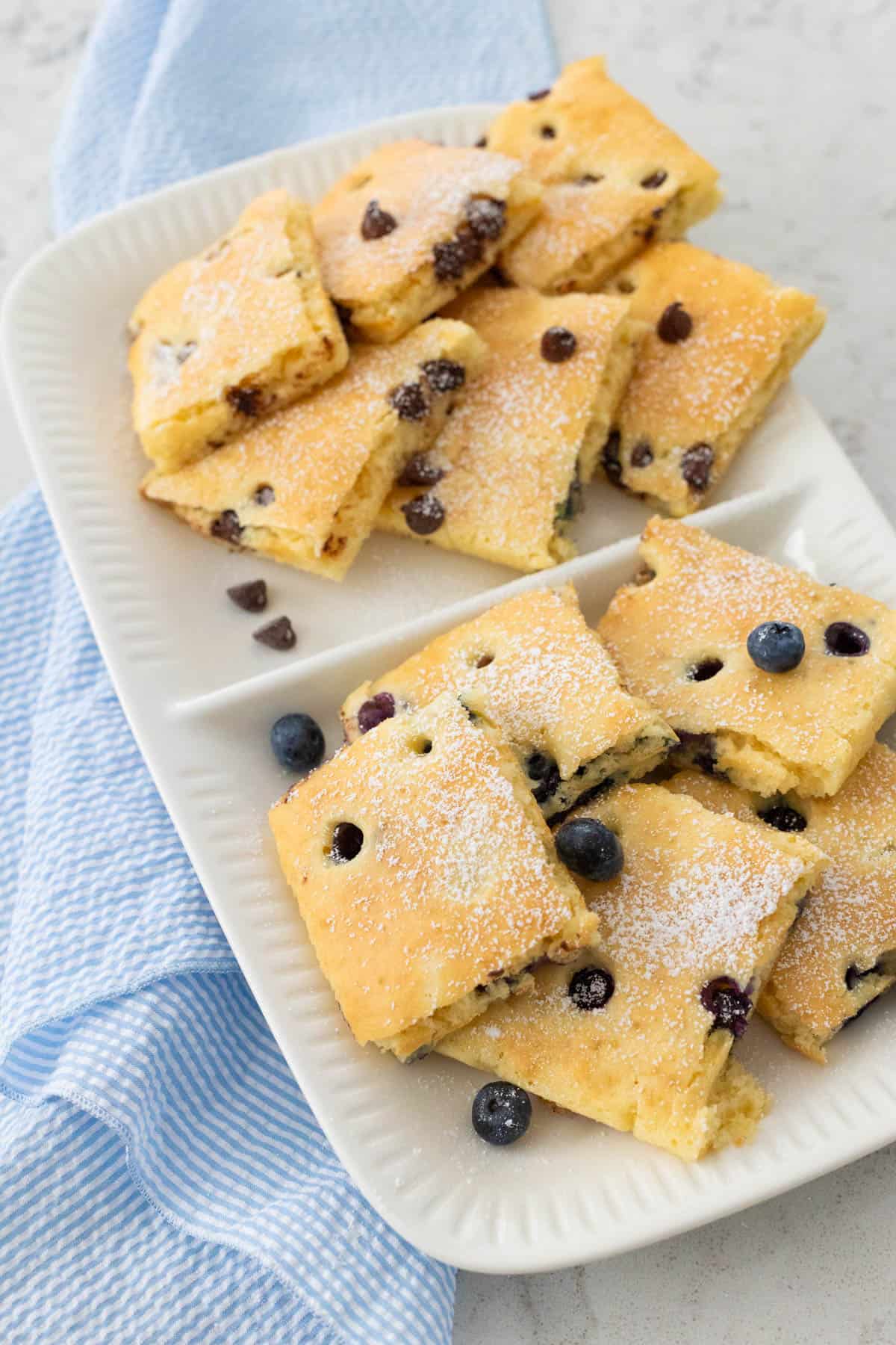 A plate of fluffy sheet pan pancakes cut into squares, topped with powdered sugar and choice of toppings.