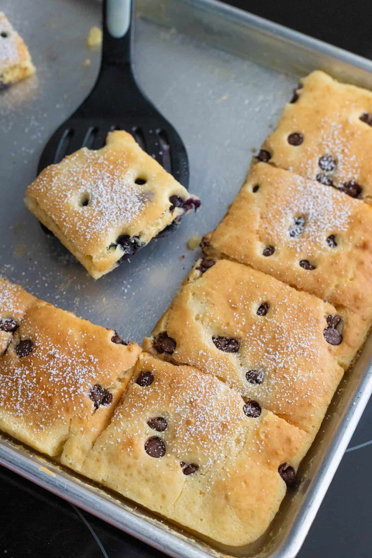 Golden sheet pan pancakes with chocolate chips, cut into squares and dusted with powdered sugar. One piece is lifted with a black spatula from a metal baking tray.