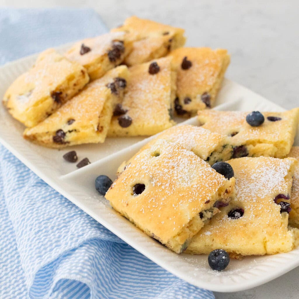 A white platter is filled with square-shaped pancakes that have been baked on a sheet pan and then sliced and served for a crowd.