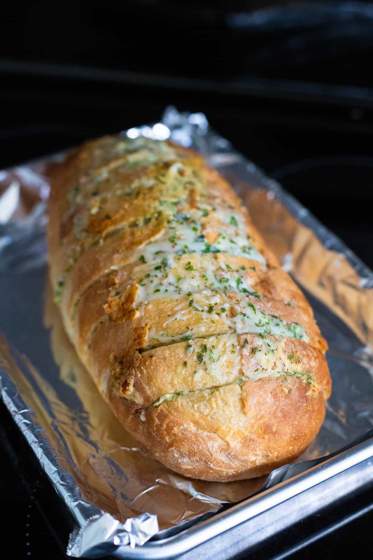 A freshly baked loaf of cheesy bread, golden and crispy with a sprinkle of parsley, sliced to reveal the melted cheese inside. The loaf is placed on a baking tray lined with aluminum foil.