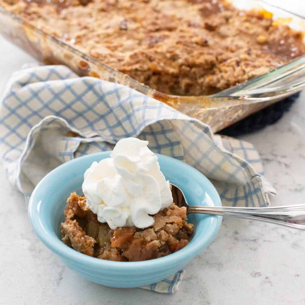 A small blue bowl is filled with some apple dump cake and topped with whipped cream.
