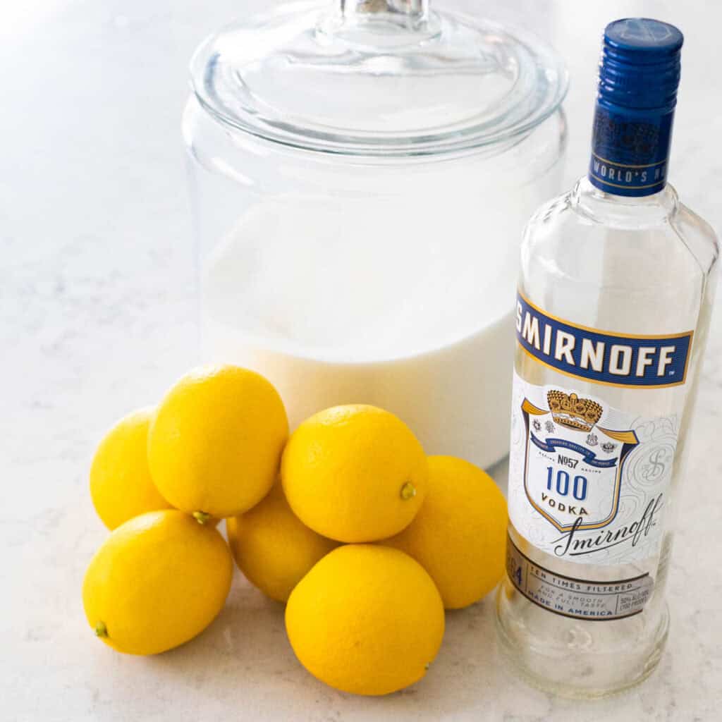 A bottle of vodka sits on the counter next to a pile of Meyer lemons and a sugar bowl.