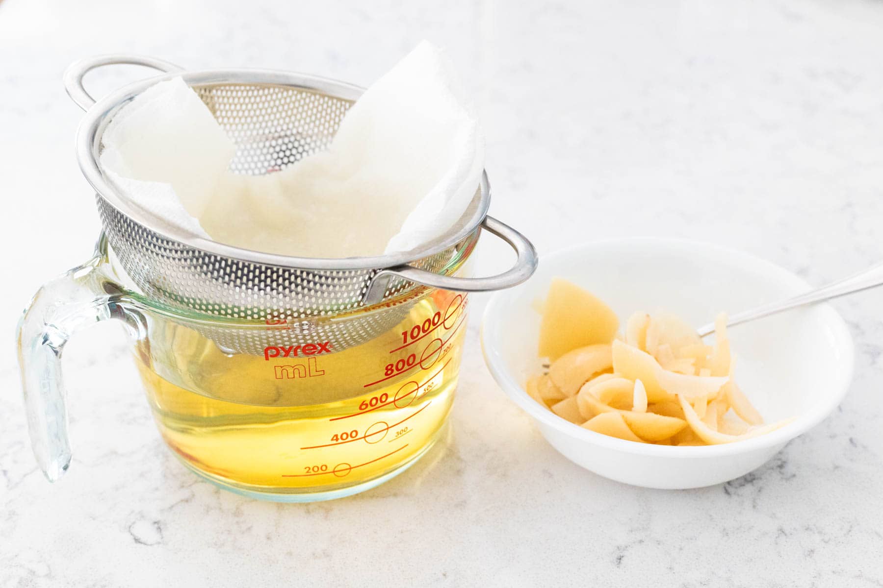 L'alcool infusé au citron étant versé à travers un tamis fin tapissé d'un filtre à papier dans un verre mesureur, avec les écorces de citron enlevées mises de côté dans un petit bol.