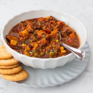 A bowl of beef and vegetable chili sits on a plate with crackers.
