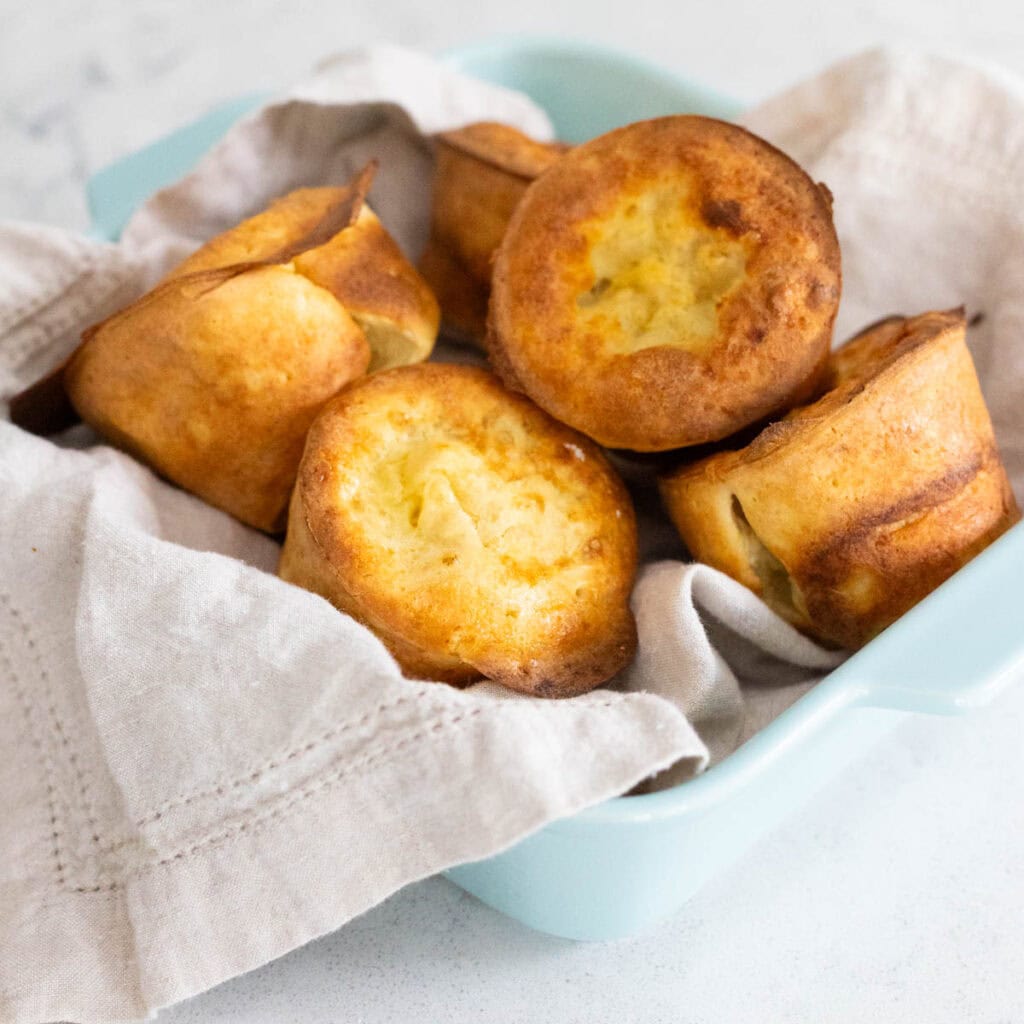 A blue dish lined with a napkin is filled with golden brown popovers.