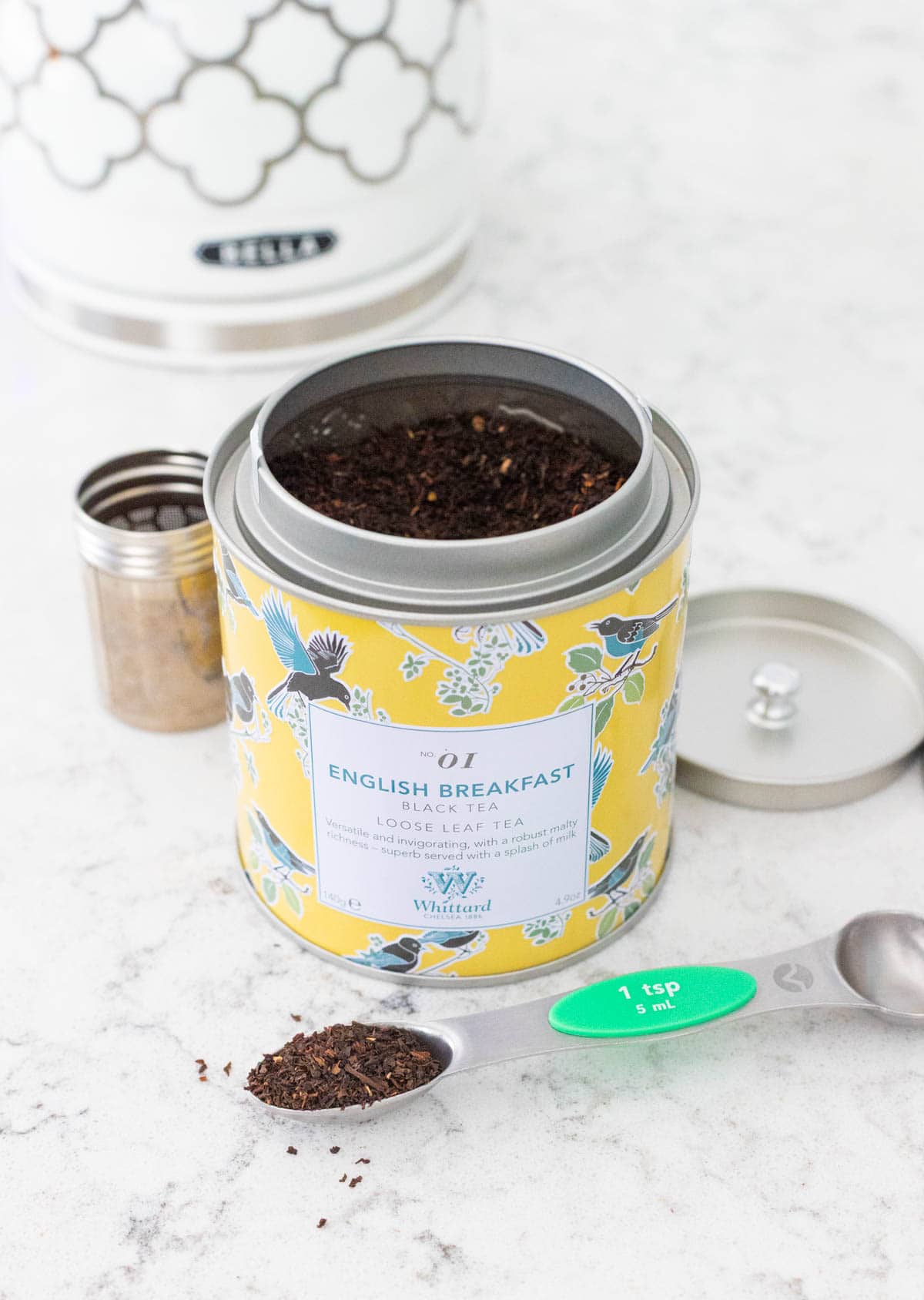 A tin of English Breakfast tea has a measuring spoon in front filled with loose leaf tea.