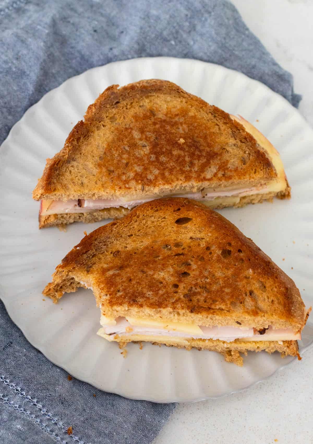 A roasted turkey and apple sandwich cut into halves on a white plate, showing melted cheese and thinly sliced turkey inside.