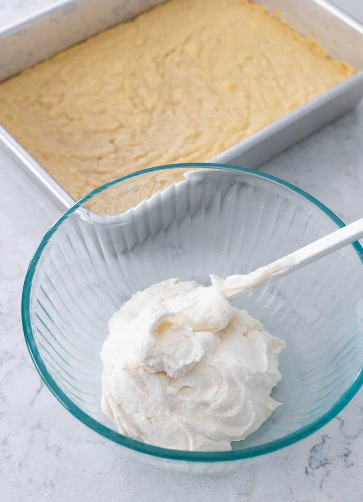 A pan of plain cookie bars sits on the counter next to a bowl of plain vanilla frosting.