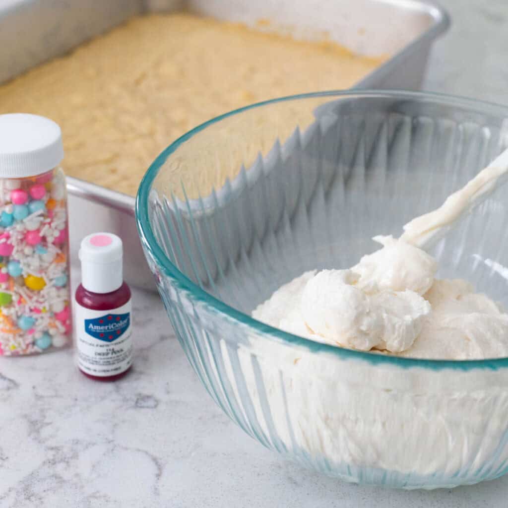 A mixing bowl is filled with vanilla frosting and it rests next to a pan of cookie bars, a jar of sprinkles, and a small bottle of food coloring gel.
