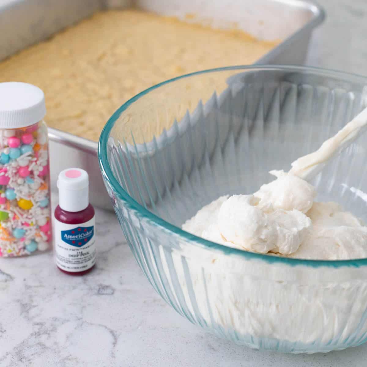 A mixing bowl is filled with vanilla frosting and it rests next to a pan of cookie bars, a jar of sprinkles, and a small bottle of food coloring gel.