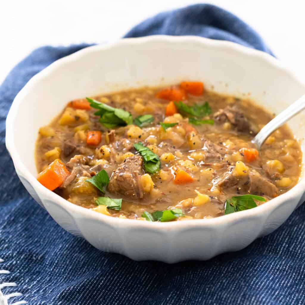 A bowl of thick, beefy stew with vegetables and barley is topped with fresh parsley.