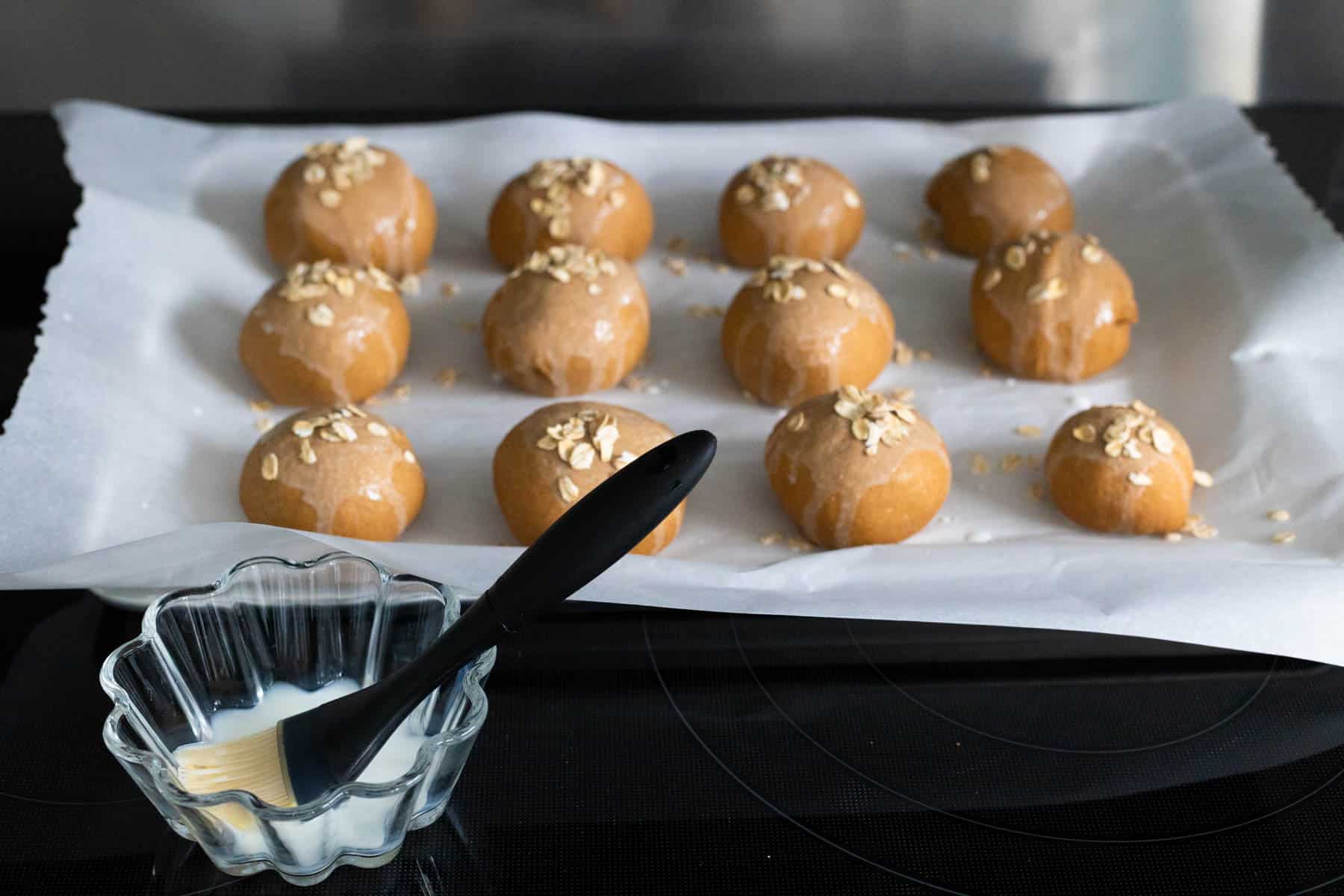 Shaped brown bread rolls on a baking sheet being brushed with milk and sprinkled with oats before baking.