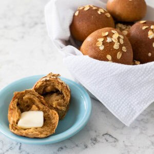A basket of brown bread rolls with oats on top sits in the back of a serving bowl with one roll broken open with melting butter on top.