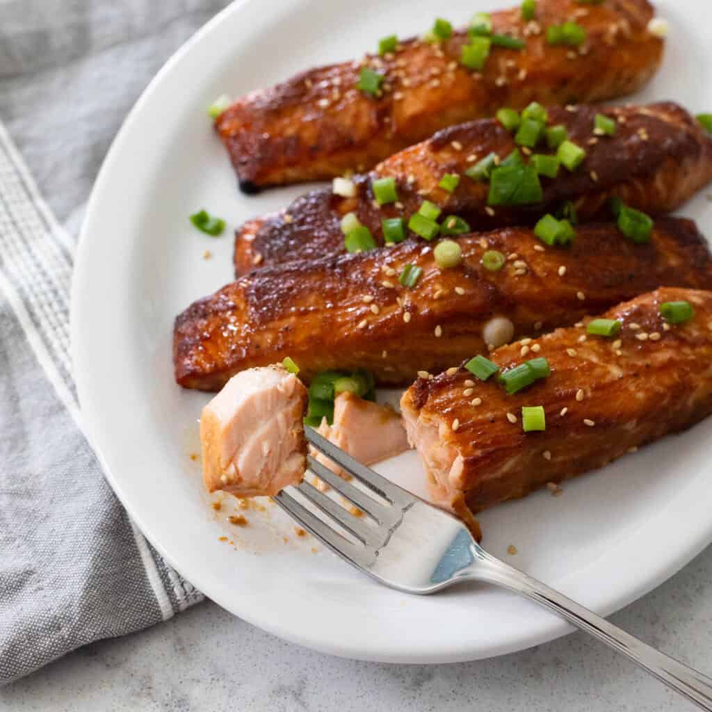 4 salmon filets have been roasted with a miso glaze and are served on a white platter. A fork takes a bite out of one.