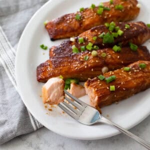 4 salmon filets have been roasted with a miso glaze and are served on a white platter. A fork takes a bite out of one.