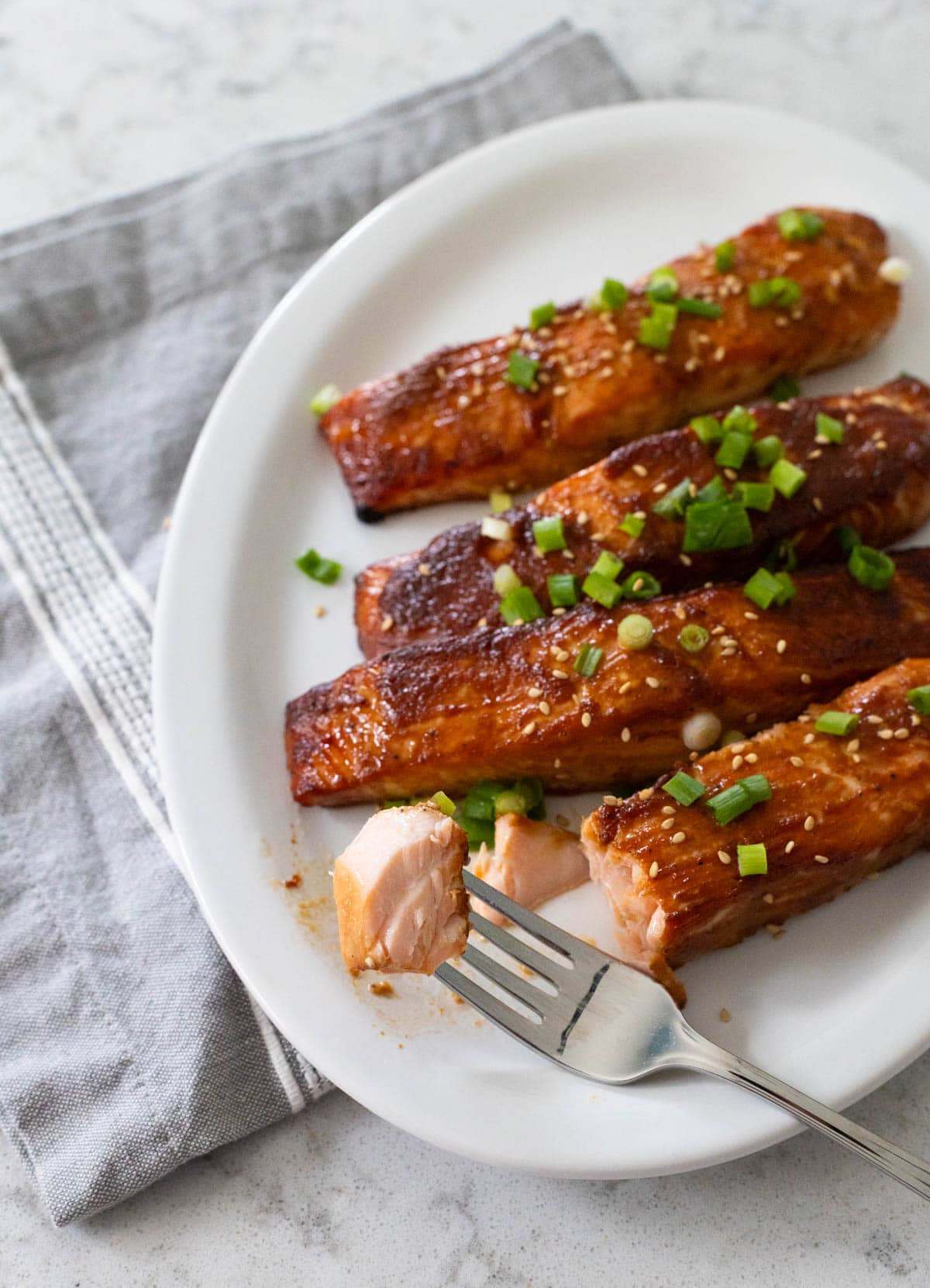4 salmon filets have been roasted with a miso glaze and are served on a white platter. A fork takes a bite out of one.