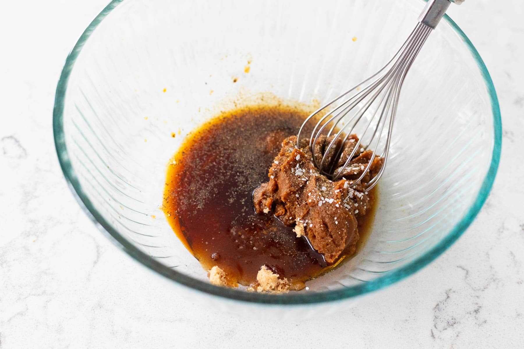 A glass bowl with miso paste, soy sauce, and seasonings being mixed with a whisk.