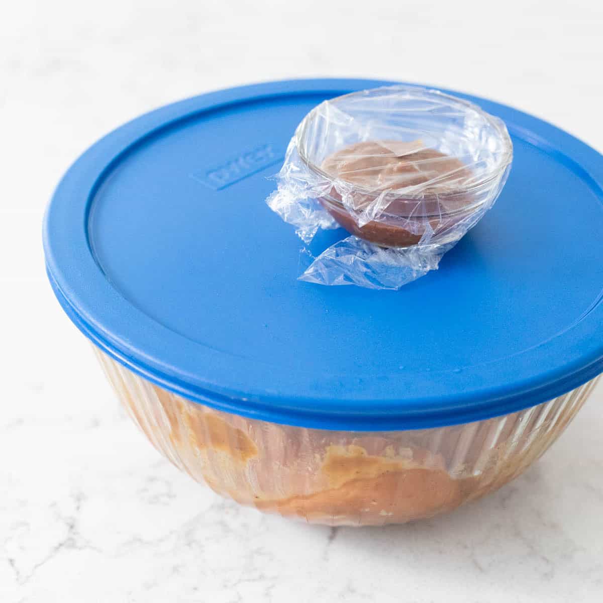 A glass bowl covered with a blue lid holding miso-marinated salmon, with extra sauce wrapped in a small bowl on top.