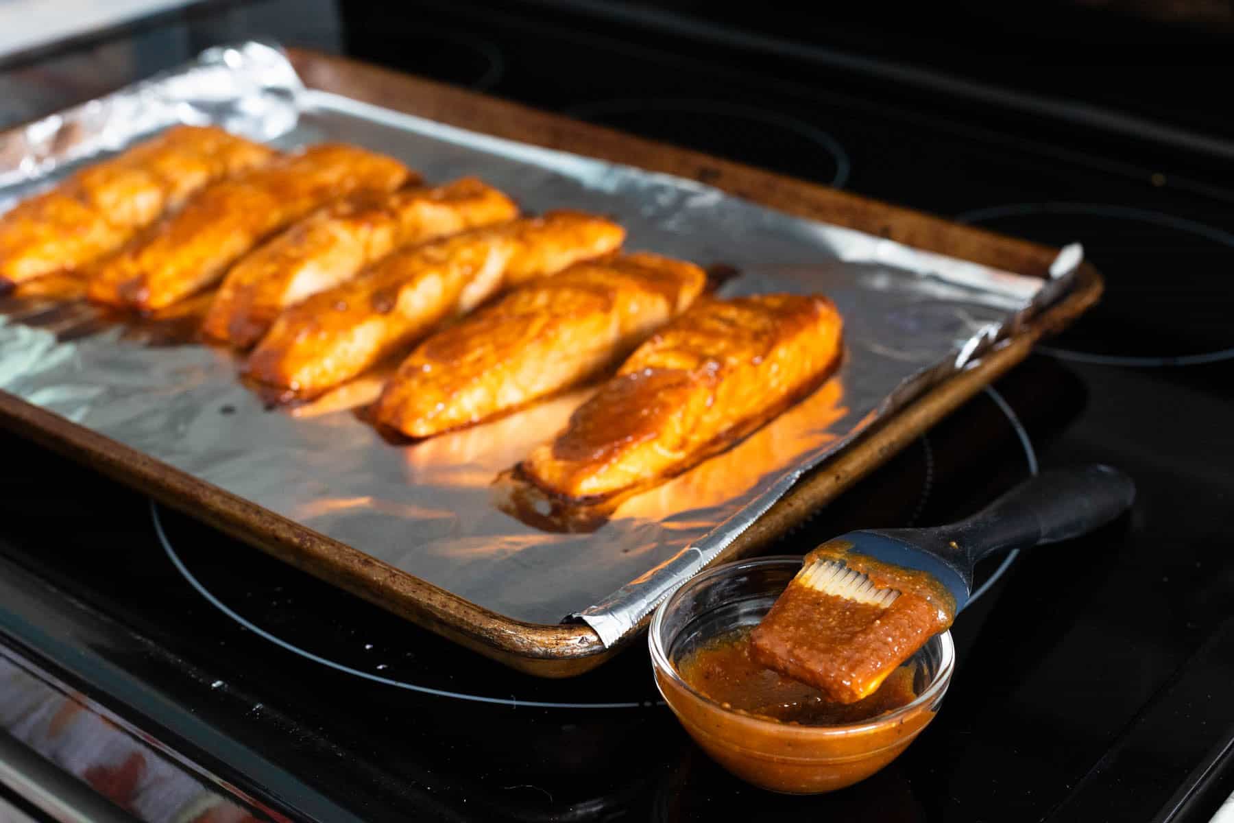 Salmon fillets on a foil-lined baking sheet being brushed with miso sauce from a small bowl.