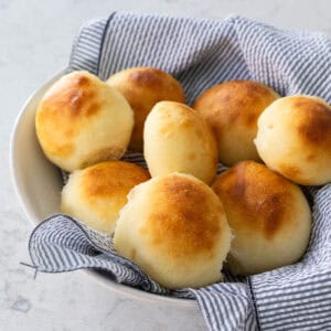 A bread basket lined with a napkin is filled with golden brown potato rolls.