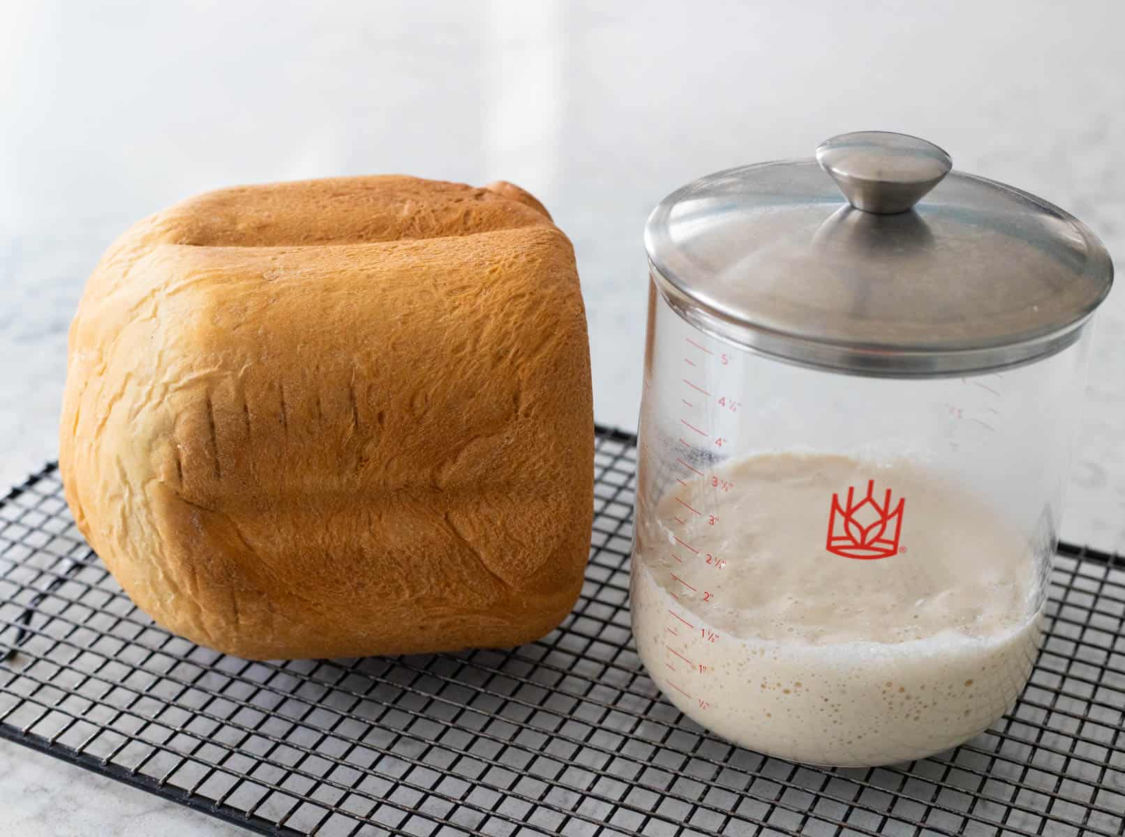 The loaf of bread is on a wire rack sitting next to a jar of sourdough starter.