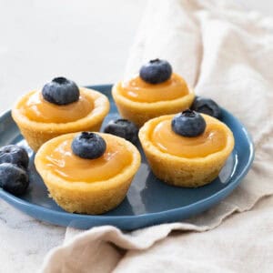 A blue plate has little lemon curd filled sugar cookie cups with fresh blueberries on top.