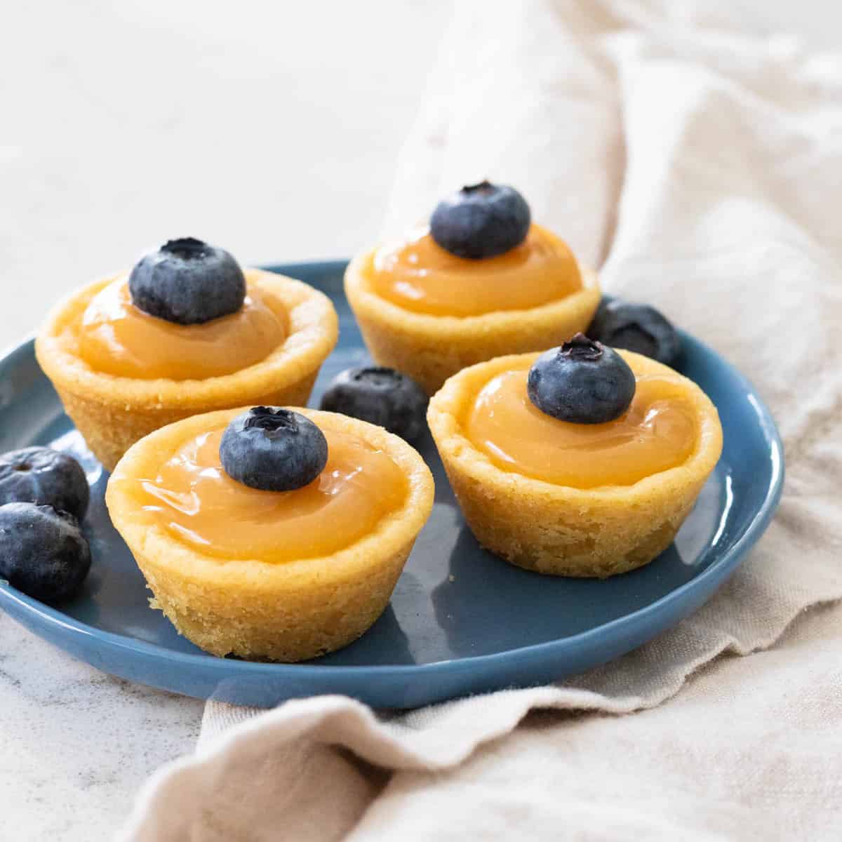 A blue plate has little lemon curd filled sugar cookie cups with fresh blueberries on top.