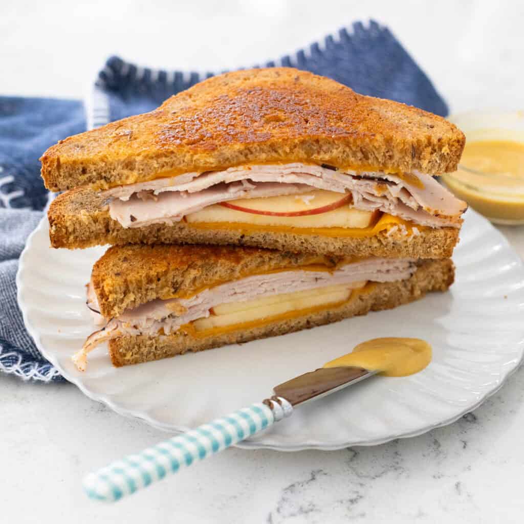 A grilled sandwich with turkey and apple is sliced open to show the fillings. A small knife with maple mustard is on the plate in front.