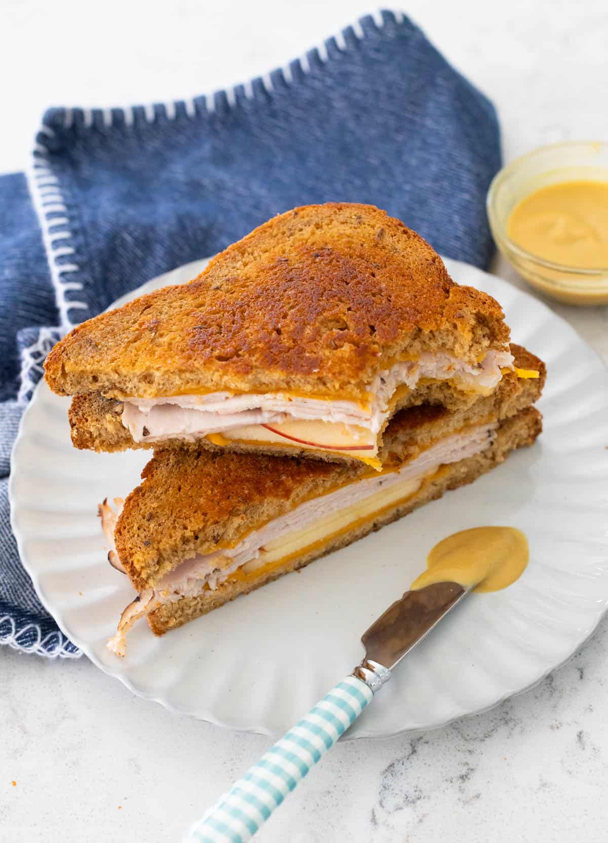 A grilled sandwich with turkey and apple is sliced open to show the fillings. A small knife with maple mustard is on the plate in front.