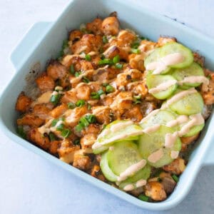 A blue baking dish has a layered baked salmon sushi rice casserole with sliced cucumber salad and a drizzle of spicy mayo on the top.