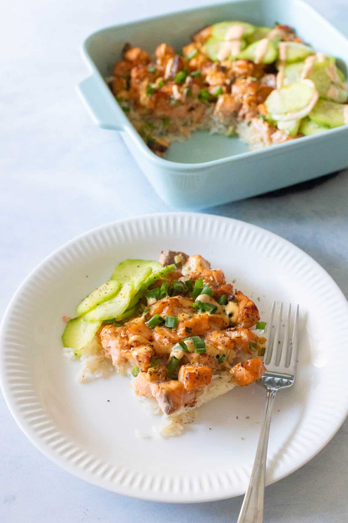 A slice of spicy salmon sushi bake topped with green onions and sliced cucumbers served on a white plate with a fork.