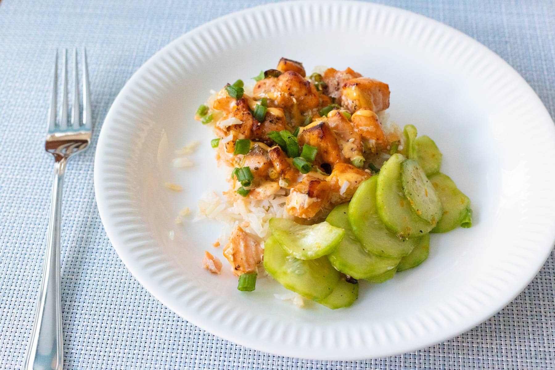 A serving of spicy salmon sushi bake over rice topped with green onions and a side of sliced cucumbers on a white plate.