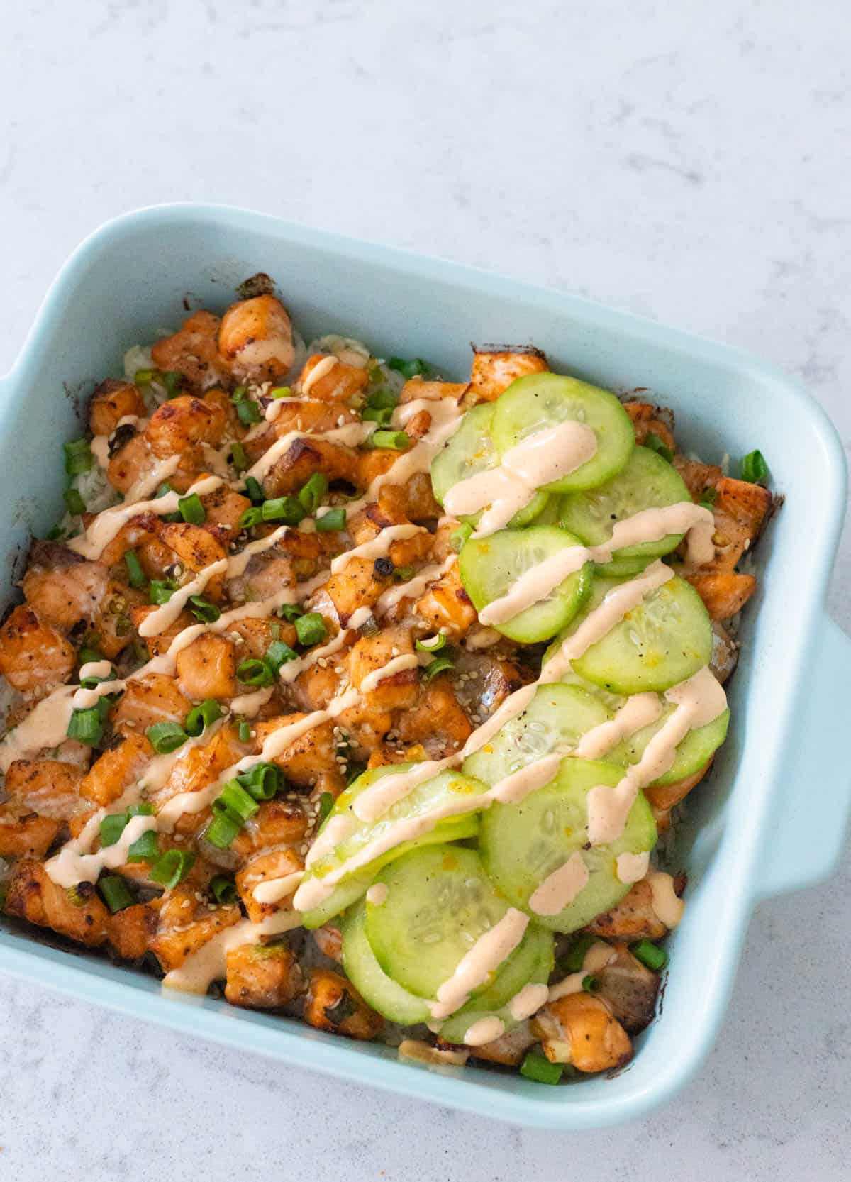 A blue baking dish has a layered baked salmon sushi rice casserole with sliced cucumber salad and a drizzle of spicy mayo on the top.